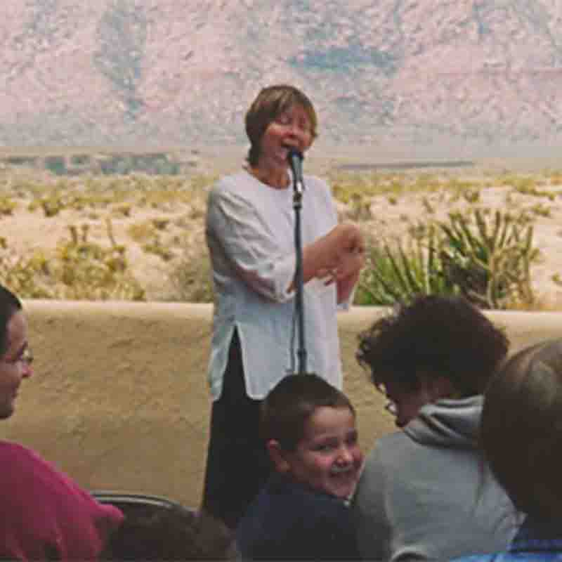 Photo of Susan Strauss gesturing and standing at a microphone in front of an audience that includes a smiling boy